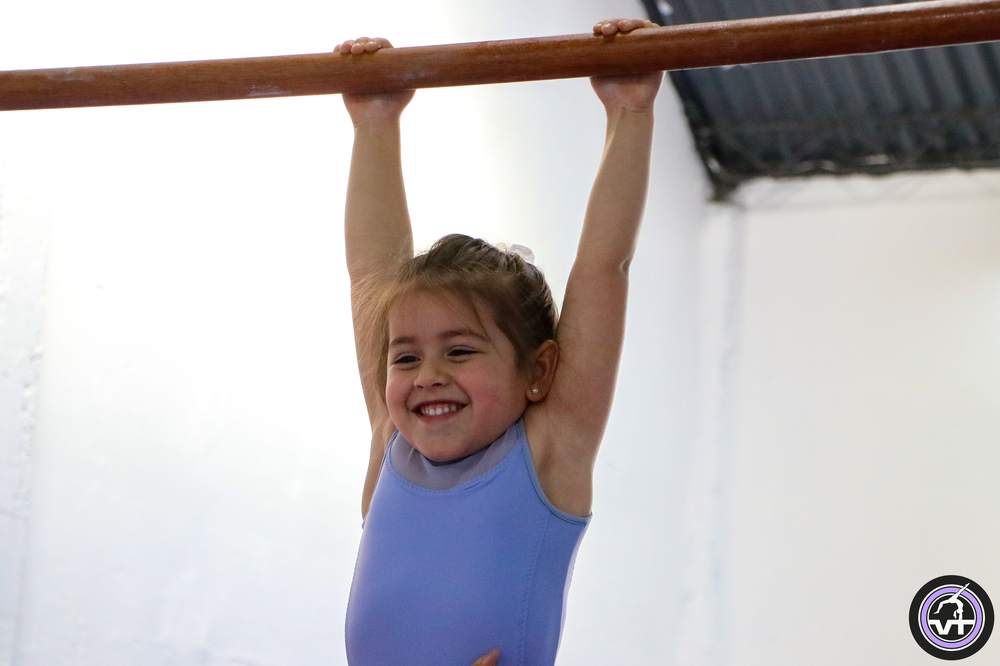Niña sonriendo colgada de la barra en una clase de gimnasia artística